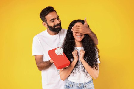 Photo for Gifts Shopping. Cheerful Middle Eastern Husband Surprising Wife With Present, Closing Her Eyes, Celebrating Her Birthday, Standing In Studio Over Yellow Background. Holiday Celebration - Royalty Free Image