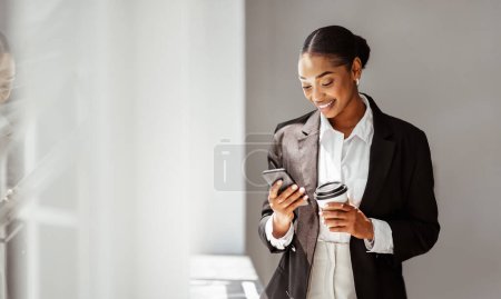 Téléchargez les photos : Joyeux jeune gestionnaire noire debout dans le bureau, boire du café pour aller et utiliser le téléphone portable, bavarder avec un partenaire d'affaires et sourire, panorama avec espace de copie - en image libre de droit