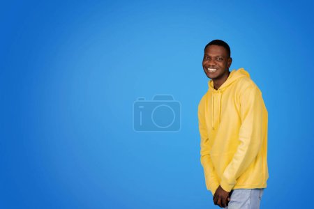 Téléchargez les photos : Gai millénial homme afro-américain en sweat-shirt s'amuse, profiter du temps libre avec de l'espace libre, isolé sur fond de studio bleu. Annonce et offre, vente, mode de vie positif et passe temps, repos et étude - en image libre de droit