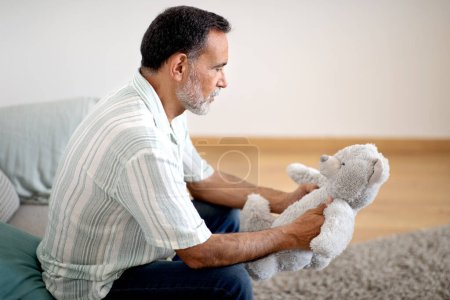 Téléchargez les photos : Dépression masculine. Homme âgé déprimé tenant un jouet, subissant une perte d'enfant ou manquant pour son enfance, assis sur le canapé à la maison. Pensionné souffrant d'un problème de pensée négative et de santé mentale - en image libre de droit