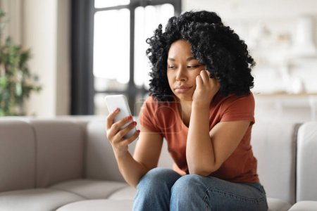 Téléchargez les photos : Jeune femme afro-américaine bouleversée assise sur le canapé et regardant l'écran du smartphone, femme noire millénaire ennuyeuse attendant un appel important ou des SMS tout en se relaxant à la maison, espace libre - en image libre de droit