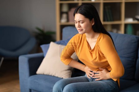 Photo for Painful Periods. Unhappy Asian Lady Struggling With Menstrual Cramps And Stomachache Pain, Touching Aching Belly Sitting On Sofa At Home. Female Health Problem, Stomach Diseases Concept - Royalty Free Image