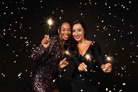 Téléchargez les photos : Joyeuses petites amies multiethniques deux jeunes femmes portant de belles tenues élégantes tenant des lumières bengales, riant parmi les éclats de confettis, célébrant ensemble le Nouvel An 2024, isolées sur fond noir - en image libre de droit