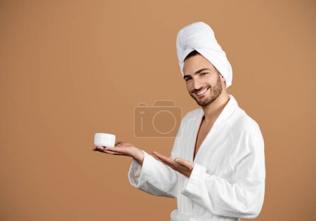 Photo for Skincare Offer. Portrait Of Hispanic Young Man Showing Moisturizing Facial Cream, Gesturing At Jar On Palm, Advertising Face Cosmetics Over Brown Studio Background, Wearing Bathrobe, Copy Space - Royalty Free Image