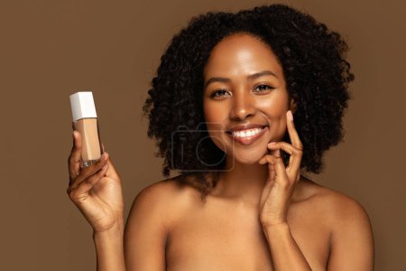 Téléchargez les photos : Souriant sexy attrayant moitié nue jeune femme noire avec une peau parfaite éclatante montrant bouteille avec fond de teint de produit de beauté ou correcteur, isolé sur fond de studio brun, gros plan - en image libre de droit