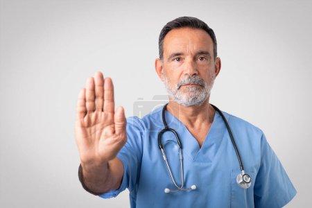 Photo for Serious confident caucasian senior doctor surgeon in blue uniform making stop sign with hand, isolated on gray background, studio. Disease control, medical care, health care, work problems - Royalty Free Image