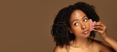 Photo for Closeup of attractive half-naked young black woman making face massage with jade gua sha in morning face care routine, looking at copy space, closeup, brown studio background, banner - Royalty Free Image