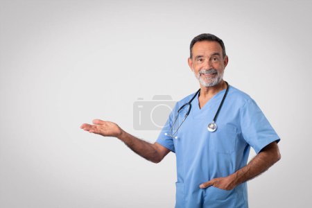 Photo for Glad caucasian senior doctor in blue uniform hold empty space on hand, isolated on gray background, studio. Recommendation medicine, approving health care, professional work advice - Royalty Free Image