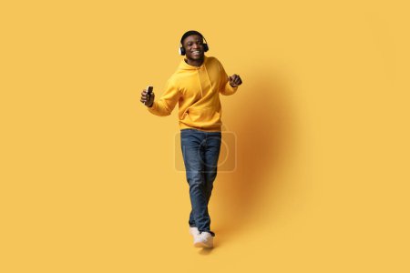Foto de Buena lista de reproducción. Feliz chico afroamericano joven guapo en casual escuchando música y bailando sobre fondo de estudio amarillo, usando auriculares inalámbricos y sonriendo, de cuerpo entero, espacio para copiar - Imagen libre de derechos