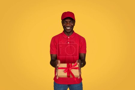 Photo for Happy millennial african american guy courier in uniform gives box gift, isolated on orange studio background. Ad and offer, home delivery, work and service, congratulations on holiday remotely - Royalty Free Image