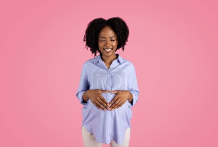 Photo for Glad young black pregnant lady in casual puts heart shape hands on big belly, enjoy love, isolated on pink studio background. Ad, offer, expect baby, future family, lifestyle - Royalty Free Image