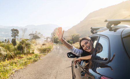 Photo for Happy pretty young woman sitting in automobile front seat, waving from car window and smiling. Family, friends, couple going summer vacation by auto together, enjoying nice weekend, sunny weather - Royalty Free Image