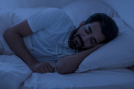 Foto de Concepto de sueño saludable. Retrato de un joven indio acostado en la cama con los ojos cerrados en la noche, tranquilo hombre del este descansando en el dormitorio, relajándose en el cuarto oscuro, teniendo hap con la mano debajo de la cabeza - Imagen libre de derechos