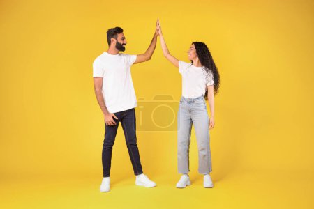 Foto de Longitud completa de la feliz pareja árabe joven dando High Five a cada uno de pie sobre el fondo del estudio amarillo, cónyuges alegres de Oriente Medio disfrutando del trabajo en equipo, celebrando el éxito común - Imagen libre de derechos