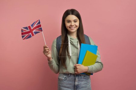 Foto de Educación en el extranjero. Chica adolescente sonriente posando con libros de trabajo y bandera británica sobre fondo de estudio rosa, estudiante femenina feliz usando mochila disfrutando aprendiendo idiomas extranjeros, espacio de copia - Imagen libre de derechos