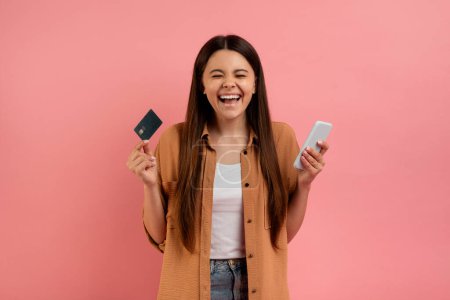 Téléchargez les photos : Mobile Shopping App. Portrait d'adolescente ravie tenant la carte de crédit et le téléphone intelligent, adolescente heureuse excitée debout sur fond de studio rose, jeune femme joyeuse faisant un achat en ligne - en image libre de droit