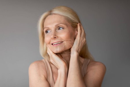 Photo for Beautiful middle aged blonde woman touching her face, attractive lady doing morning face care routine, grey wall background, closeup portrait, copy space - Royalty Free Image