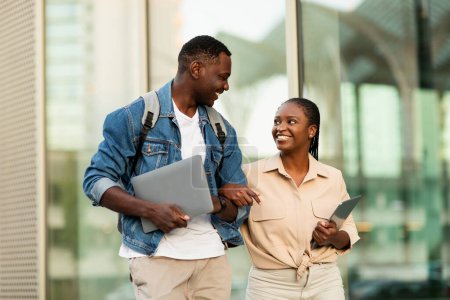 Foto de Alegre joven negro hombre y mujer estudiantes yendo a la universidad juntos, sosteniendo gadgets en sus manos, llevando mochilas, pareja caminando por la calle, tener conversación y sonriendo - Imagen libre de derechos