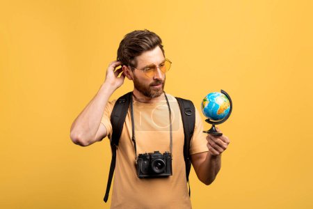 Téléchargez les photos : Voyageur masculin d'âge moyen réfléchi tenant un petit globe, regardant et pensant où aller ensuite, debout avec un sac à dos et un appareil photo sur fond de studio jaune - en image libre de droit