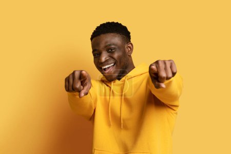 Photo for Positive cheerful cool handsome millennial african american guy wearing yellow hoodie gesturing, pointing with both hands and smiling at camera, isolated on yellow studio background - Royalty Free Image