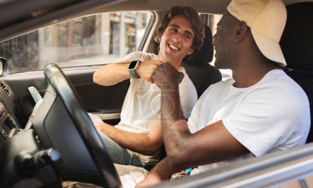 Téléchargez les photos : Cool heureux jeunes gars multiethniques portant tenue décontractée assis à l'intérieur de l'auto, se donnant une bosse poing et souriant, se préparer pour la promenade en voiture, profiter week-end voyage ensemble - en image libre de droit