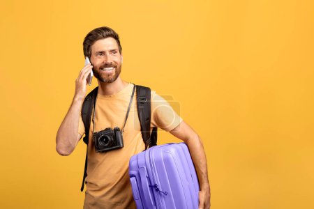 Téléchargez les photos : Communication de voyage. Homme d'âge moyen souriant parlant sur son téléphone portable, tenant sa valise et regardant de côté l'espace libre, attendant son vol et communiquant, fond jaune - en image libre de droit