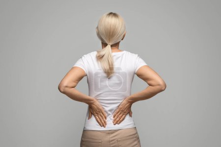 Unrecognizable Blonde Woman Suffering Body Ache Or Kidneys Pain, Sick Mature Female Presseing Her Hands To Lower Back, Massaging Inflamed Area, Standing Over Gey Studio Background, Copy Space