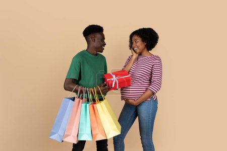 Photo for Cheerful millennial black husband with many packages gives box to wife, enjoy pregnancy, isolated on beige background, studio. Family lifestyle, expecting baby, celebration and surprise, shopping - Royalty Free Image