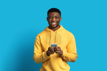 Téléchargez les photos : Joyeux beau jeune homme noir cool en sweat à capuche jaune à l'aide d'un smartphone et souriant, isolé sur fond de couleur bleue, espace de copie. Africain américain gars vérifier la plus récente application mobile, bavarder - en image libre de droit