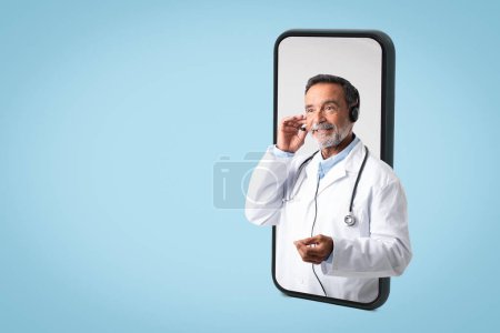 Photo for Happy caucasian senior doctor therapist in white coat, headphones consults client remotely on large phone screen, isolated on blue background, studio. Support service, medical care, health care - Royalty Free Image