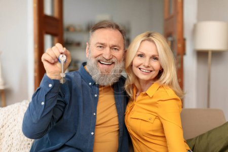 Photo for Mortgage For Seniors. Joyful mature couple moving into new home, man showing key and embracing woman in modern living room, smiling to camera, Happy senior house buyers. Real estate concept - Royalty Free Image