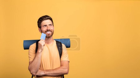 Photo for Travel budget concept. European middle aged man with tourist equipment holding credit card and thinking, planning funds for his camping adventure over yellow studio background, free space - Royalty Free Image