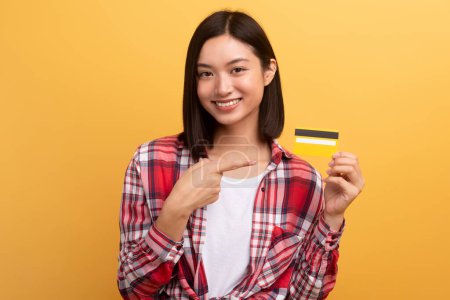 Photo for Cheerful japanese lady shopaholic pointing finger at credit card, recommending sale isolated on yellow background. Finance and money for shopping, ad and offer - Royalty Free Image