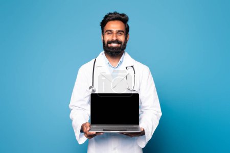 Photo for Happy hindu man doctor in white coat with stethoscope showing laptop with blank screen on blue studio background. Professional work, medical care, recommendation, ad and offer - Royalty Free Image
