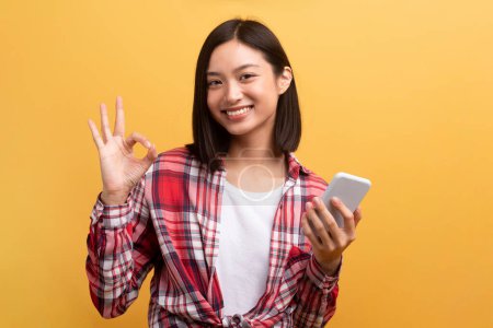 Téléchargez les photos : Sourire jeune femme coréenne tenant smartphone et montrant geste ok, recommander une nouvelle application ou un site Web, démontrant signe ok sur fond jaune - en image libre de droit