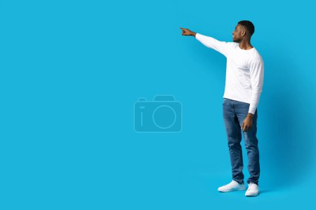 Photo for Handsome black guy in casual outfit pointing at copy space for text, advertisement over blue studio background, touching something invisible, blank space for gadget screen, full length - Royalty Free Image