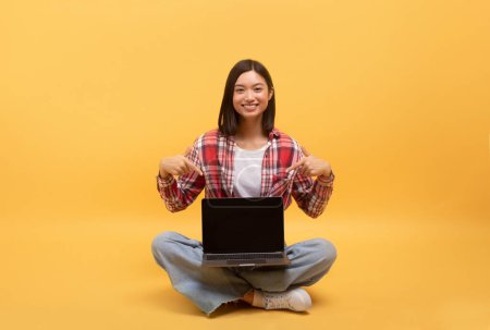Photo for Happy asian lady in casual pointing fingers at laptop with black screen, sitting on floor on yellow background, mockup. Blogger recommendation and device for work, study - Royalty Free Image