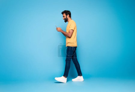 Téléchargez les photos : Indien en tenue décontractée marchant avec smartphone et souriant sur fond de studio bleu, en utilisant une application mobile agréable, pleine longueur, vue latérale, espace de copie pour l'annonce - en image libre de droit