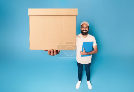 Téléchargez les photos : Livreur indien tenant boîte de carton colis étirant à la caméra, debout sur fond bleu et souriant, au-dessus de la vue. Transport et prestation de services - en image libre de droit