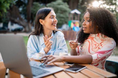 Photo for Cheerful Female Friends Shopping Online On Laptop While Resting In Cafe Outdoors, Two Happy Multiethnic Women Pointing At Screen And Discussing Purchases, Sitting At Table Outside And Drinking Coffee - Royalty Free Image