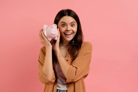 Photo for Funny cute teen girl shaking piggy bank, listening sound of coins, checking saved money, happy excited female teenager enjoying economy, standing over pink studio background, copy space - Royalty Free Image