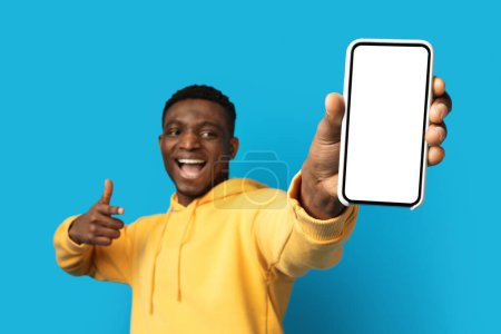 Téléchargez les photos : Cool émotionnel jeune homme afro-américain en sweat à capuche jaune montrant grand smartphone avec écran blanc vide et geste pouce levé, isolé sur fond bleu, recommander belle application mobile - en image libre de droit