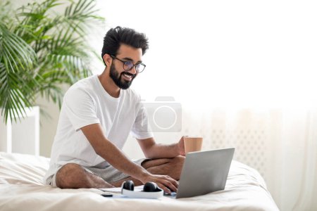 Photo for Handsome Young Indian Man Using Laptop And Drinking Coffee While Sitting In Bed, Happy Eastern Man Browsing Internet On Computer And Enjoying Hot Drink While Relaxing In Cozy Bedroom, Copy Space - Royalty Free Image