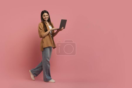 Foto de Sonriente chica adolescente con el ordenador portátil en las manos caminando sobre fondo rosa, tiro de longitud completa de hermosa adolescente femenino utilizando la computadora para el estudio en línea, Disfrutando de la enseñanza a distancia, Copiar el espacio - Imagen libre de derechos