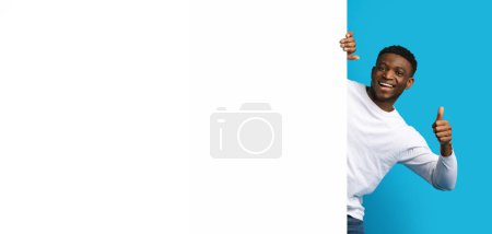 Téléchargez les photos : Positif heureux beau jeune homme noir posant à côté du tableau publicitaire blanc blanc horizontal, montrant pouce levé et souriant, fond bleu, maquette, espace vide, bannière. Grande offre, affaire - en image libre de droit