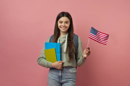 Téléchargez les photos : Joyeux adolescent échange étudiant fille avec des ordinateurs portables tenant Etats-Unis drapeau américain, joyeuse adolescente étudier l'anglais à l'étranger, regardant et souriant à la caméra, posant sur fond de studio rose, espace de copie - en image libre de droit