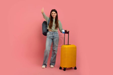Photo for Teen Travel Programs. Positive Young Girl With Tickets And Suitcase Showing Thumb Up Gesture While Posing On Pink Studio Background, Cute Female Teenager Enjoying Travelling, Full Length, Copy Space - Royalty Free Image