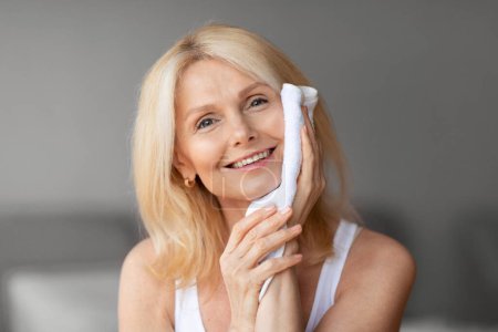 Photo for Daily hygiene. Beautiful middle aged woman wiping face with towel, making morning routine, sitting in bedroom and smiling at camera at home, copy space - Royalty Free Image