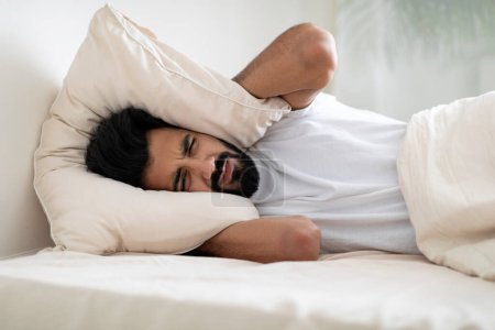 Photo for I Cant Sleep. Portrait of irritated young indian man lying in bed and covering ears with pillow, hearing and suffering from too loud sound, angry millennial eastern guy tired of noisy neighbors - Royalty Free Image