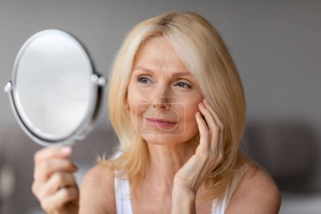 Photo for Beautiful middle aged blonde woman touching face and looking in mirror, enjoying her flawless skin, smiling female making anti-aging beauty routine, closeup - Royalty Free Image